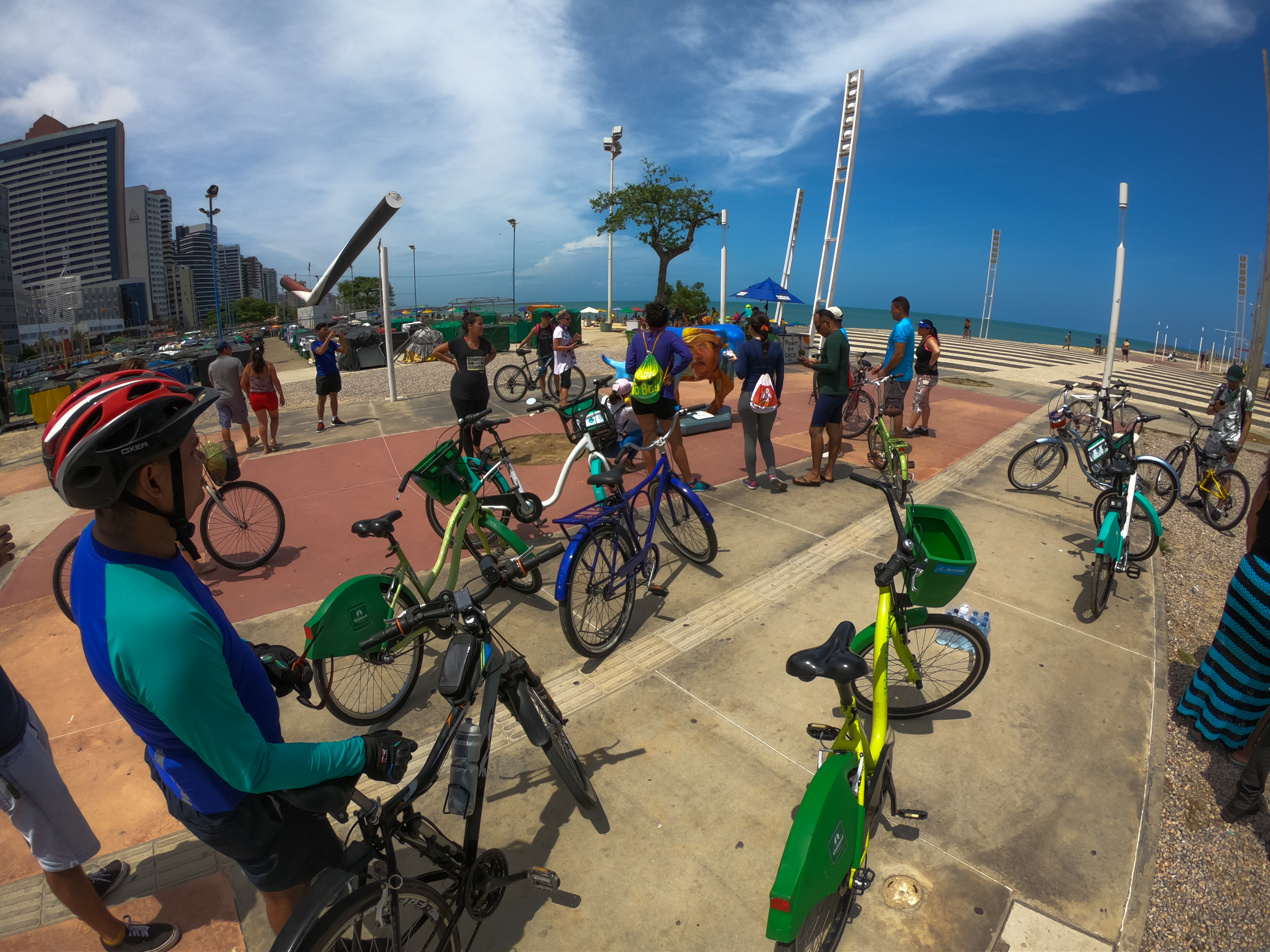 Ciclistas com bicicletas paradas em cima de calçada com o mar e espigão da Beira-Mar ao fundo